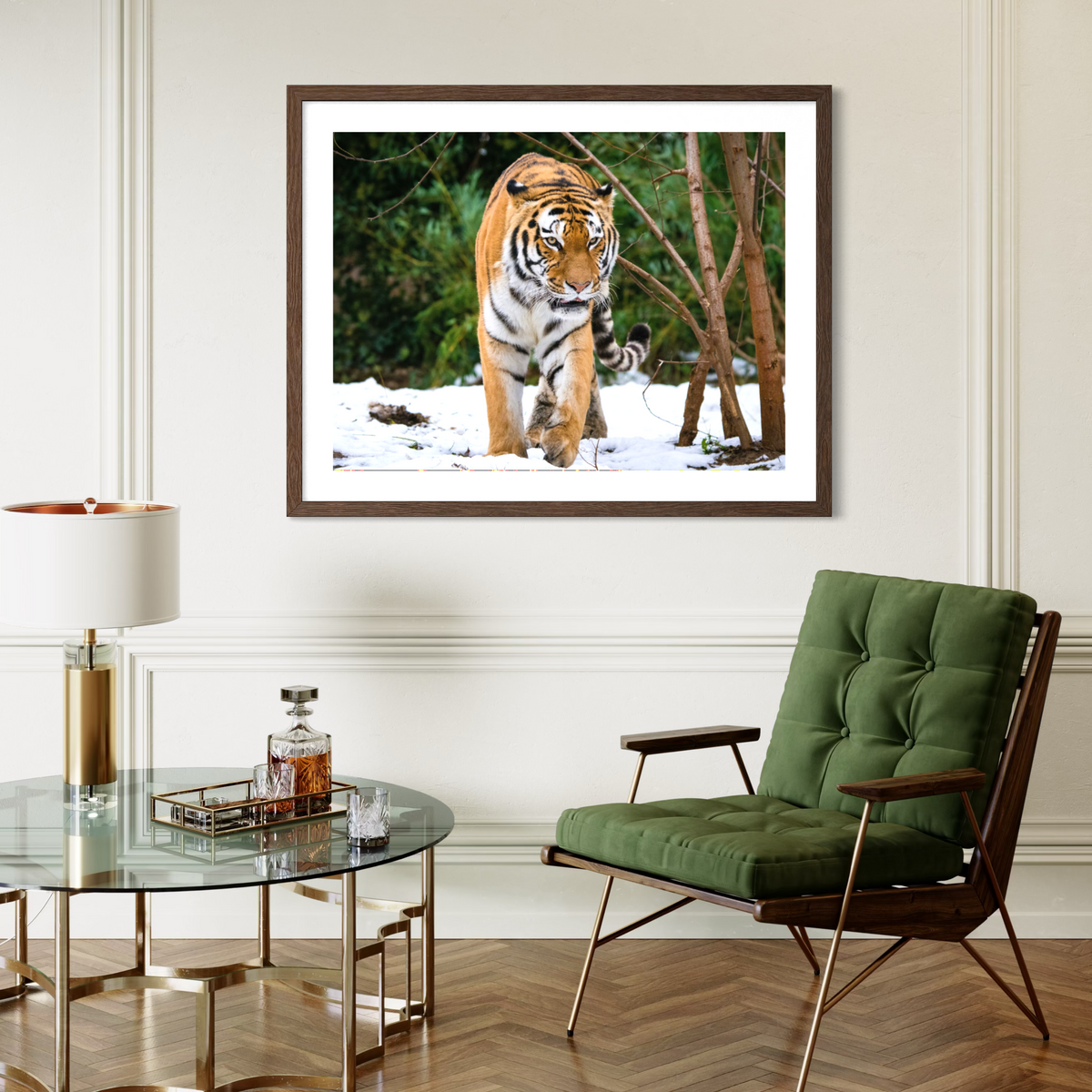 A framed dark wood wall art print depicting a tiger walking through snow and trees, displayed in a modern living room setting above a green armchair and next to a glass coffee table.