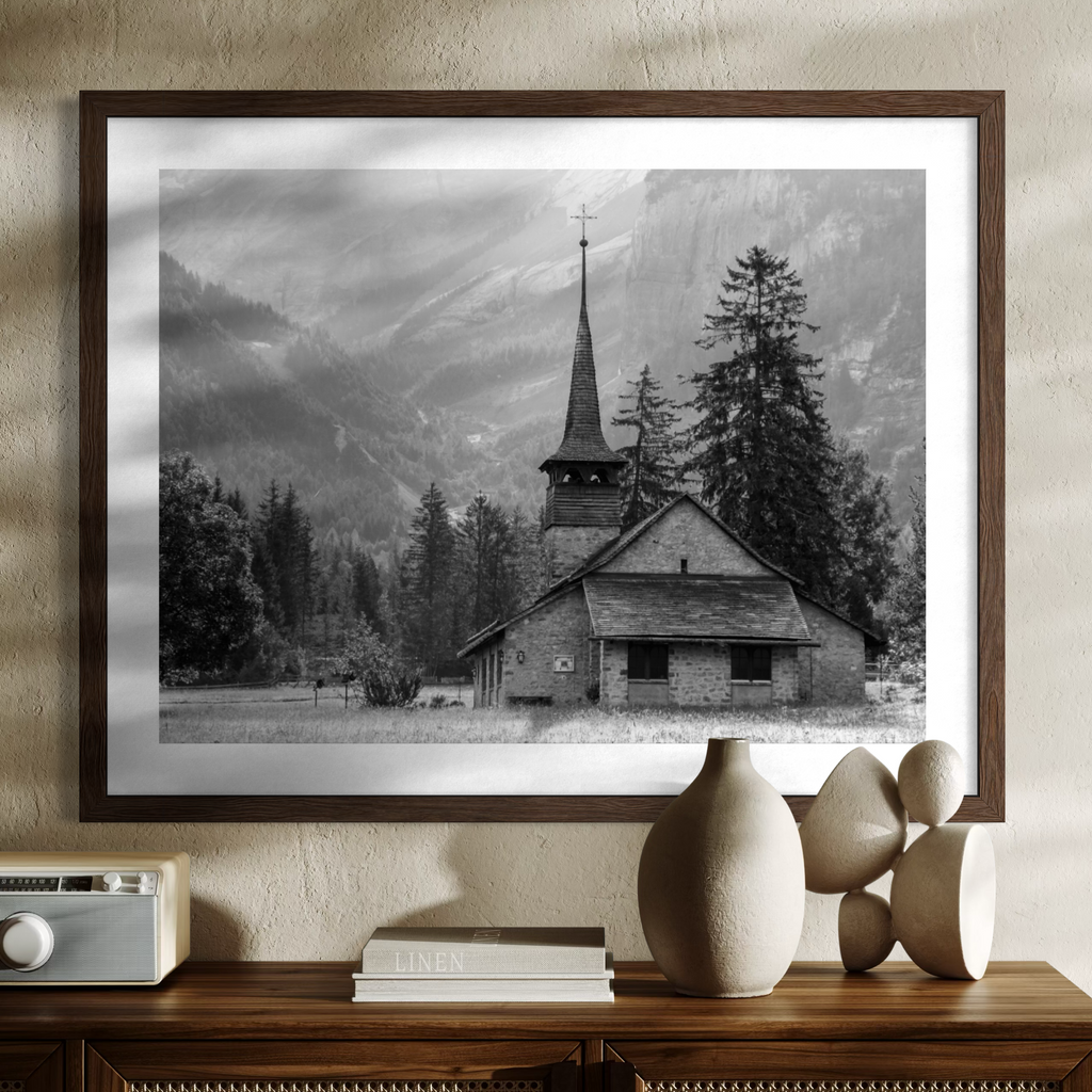 A framed black and white landscape photograph of a mountain church and pine trees, displayed on a textured beige wall above a wooden console table with decorative objects.