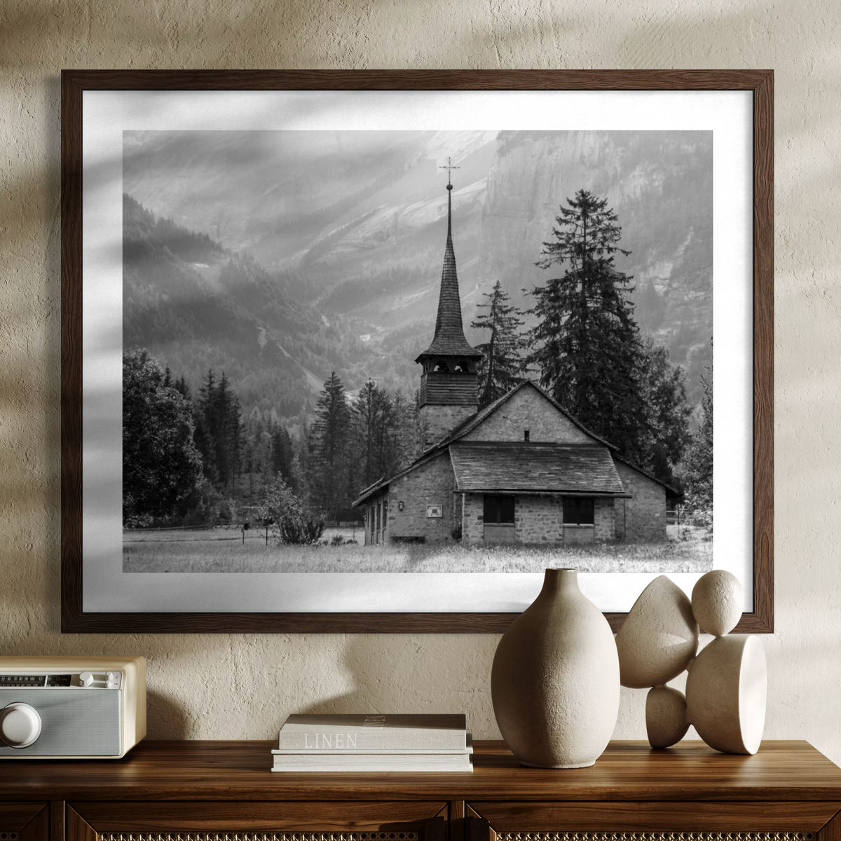 A framed black and white landscape photograph of a mountain church and pine trees, displayed on a textured beige wall above a wooden console table with decorative objects.