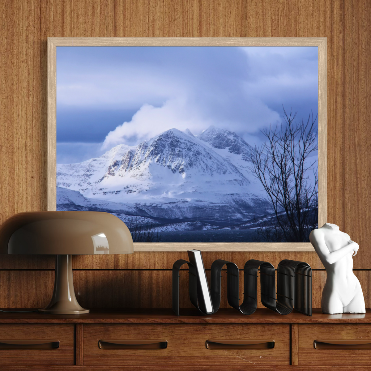 A framed wall art print depicting a snowy mountain landscape under a cloudy blue sky, displayed above a wooden console table with a lamp, books, and a white figurine.