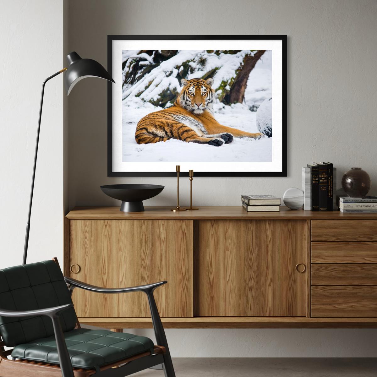 A black framed art print of a tiger lying in the snow, displayed on a light gray wall above a wooden credenza in a modern living room setting.